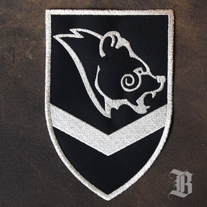 Puede incluir: Parche bordado en negro y plata con un diseño de cabeza de oso. El parche tiene forma de escudo con un galón y borde plateados. La cabeza del oso es blanca con detalles. El fondo es negro.