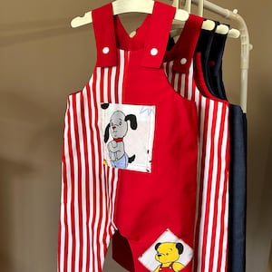 May include: A red overall with white vertical stripes on one side and a solid red panel on the other. It features a square chest pocket with a cartoon dog illustration and a diamond-shaped patch on the leg depicting a yellow bear character.