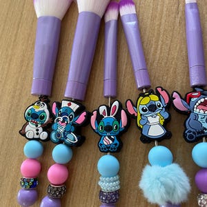May include: A collection of six makeup brushes with purple handles and white and purple bristles. Each brush has a different cartoon character charm, with blue, pink, and purple beads. The brushes are displayed on a light brown surface.