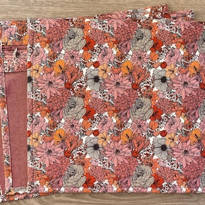 May include: Floral pattern placemats in shades of pink, orange, and gray. The placemats feature a repeating design of stylized flowers on a white background. The edges of the placemats are a solid burgundy color.