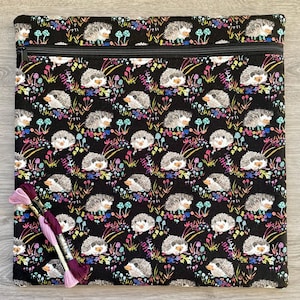 May include: A black zippered pouch with a colourful print of hedgehogs and mushrooms. The pouch is made of fabric and has a zipper closure. Two spools of embroidery floss are in front of the pouch.