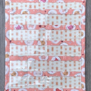 May include: A quilted organiser with clear pockets and a cat-themed fabric design. The fabric features repeating cat faces in shades of peach, pink, and cream, with a dotted background. The edges are trimmed with a peach border.