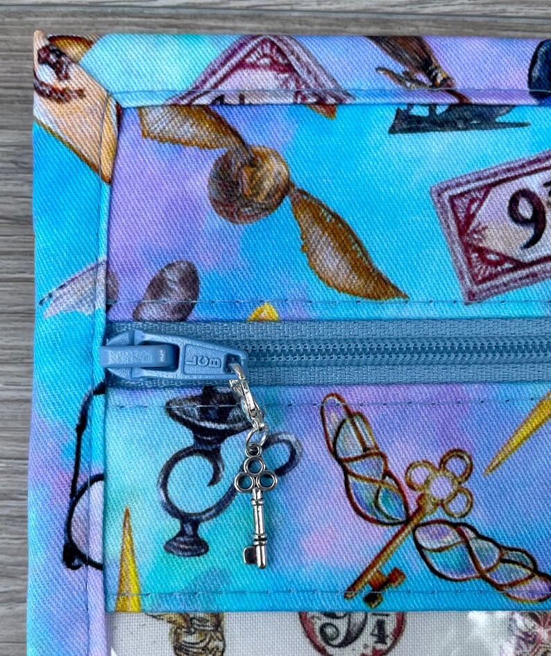 Może przedstawiać: Niebieska, materiałowa kosmetyczka zapinana na zamek błyskawiczny z kolorowym nadrukiem w stylu Harry'ego Pottera. Suwak ma srebrny brelok w kształcie klucza.