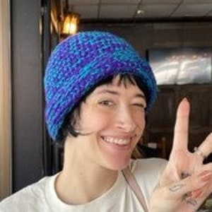 May include: A woman wearing a blue and purple knitted beanie with a white t-shirt and giving a peace sign.