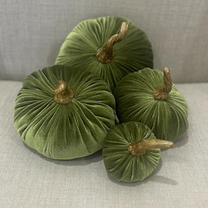 May include: Four green velvet pumpkins with gold stems. The pumpkins are arranged in a pyramid shape on a light gray surface.