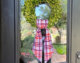 May include: A green boxwood wreath with a red and white plaid bow hanging on a glass door.