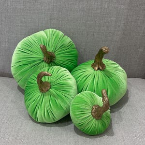 May include: Four green velvet pumpkins with gold stems. The pumpkins are arranged in a pyramid shape, with the largest pumpkin at the bottom and the smallest pumpkin at the top.