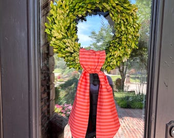 May include: A green boxwood wreath with a pink and white striped bow. The wreath is hanging on a dark brown door.