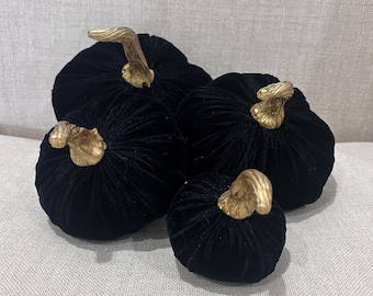 May include: Four black velvet pumpkins with gold stems. The pumpkins are arranged in a group, with the largest pumpkin in the back and the smallest pumpkin in the front.