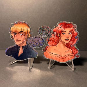 May include: Two holographic stickers of stylized characters. One has blonde hair and wears a dark blue top. The other has red hair and an orange top. Speech bubbles with an apple and a wolf are included.