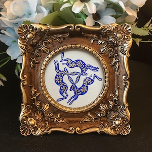 May include: A square, ornate, metallic gold-toned frame with intricate scrollwork and floral corner details. Inside the circular centre, a white background features a blue and gold illustration of three hares arranged in a circular pattern.