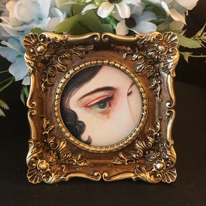 May include: A close-up of a framed painting of a woman's eye with green iris and brown eyelashes. The frame is gold with ornate details.