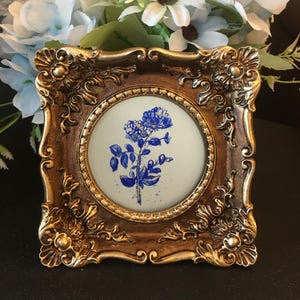 Może przedstawiać: Kwadratowa, ozdobna, złota rama do zdjęć z okrągłym, kremowym środkiem. Środek przedstawia niebieski kwiatowy obraz akwarelowy. Rama ma szczegółowe zdobienia i estetykę vintage, odpowiednią do dekoracji wnętrz.