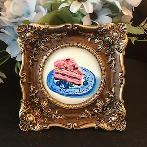 Puede incluir: Una pintura enmarcada de una rebanada de pastel rosa con chispas en un plato azul y blanco.
