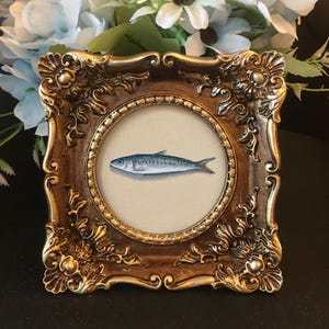 Può includere: Piccolo dipinto incorniciato di un pesce. L'opera d'arte è esposta in un'apertura rotonda, racchiusa da una cornice dorata ornata. Il pesce è blu e argento, dipinto su uno sfondo beige.