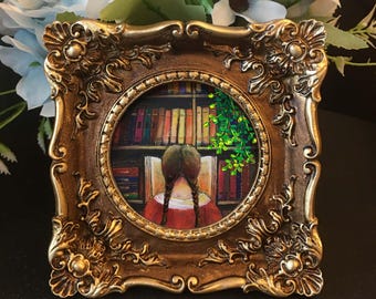 May include: A small, ornate, gold-colored picture frame with a round interior. The framed artwork depicts a person with braided hair reading in front of a bookshelf. Green foliage hangs from the top right corner.