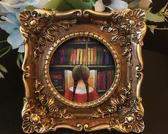 May include: A framed miniature painting depicts a person with braided hair reading in front of a bookshelf. The painting is set within a gold-colored, ornate frame. The artwork features a warm color palette with reds, browns, and yellows.