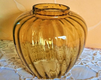 May include: A clear glass vase with a ribbed design. The vase is amber in color and has a round shape. It is sitting on a white crocheted tablecloth.