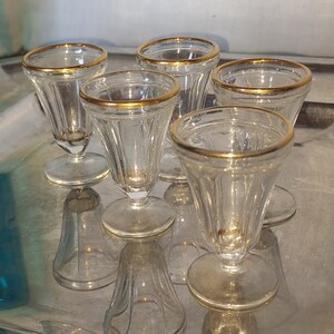 Peut inclure: Cinq verres à shot en verre transparent avec des bords dorés sont disposés sur un plateau argenté. Les verres sont disposés en demi-cercle.