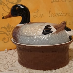 Peut inclure: Un beurrier en céramique en forme de canard avec un couvercle brun. Le canard est peint avec des détails réalistes, y compris des yeux noirs, un bec noir et des plumes brunes et blanches.