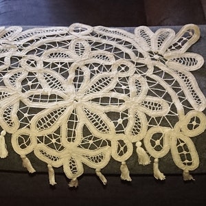 Op de afbeelding: Een wit gehaakt kanten tafelkleed met een gegolfde rand en franjes. De kant heeft een delicaat bloemenpatroon.