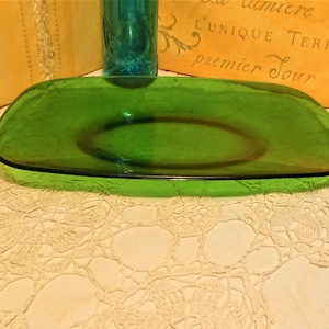 Peut inclure: Un plat de service en verre vert avec un bord légèrement surélevé. Le plat est posé sur une dentelle blanche au crochet.