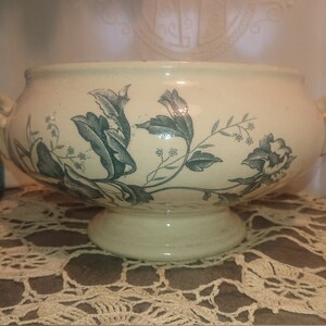 Peut inclure: Un bol de service en céramique blanche avec un motif floral vert. Le bol a deux poignées et un piédestal.
