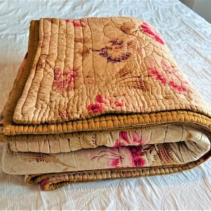 Könnte beinhalten: Eine gefaltete Steppdecke mit floralem Muster und goldfarbenem Besatz. Die Steppdecke ist aus einem weichen, leichten Stoff gefertigt und perfekt, um jedem Schlafzimmer einen Hauch von Vintage-Charme zu verleihen.