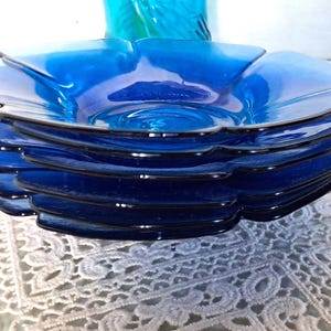May include: A stack of six cobalt blue glass bowls. Each bowl has a petal-like rim and a slightly concave center. The bowls are stacked on a white lace tablecloth, with a turquoise glass object in the background.