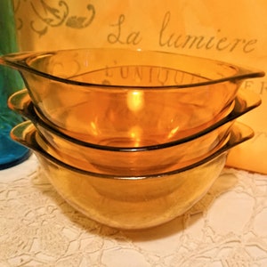May include: Three amber glass bowls stacked on top of each other. The bowls are round and have a slight lip around the top edge. The bowls are sitting on a white crocheted tablecloth.
