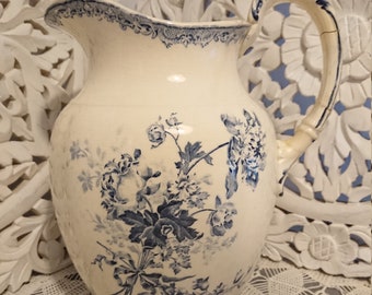 May include: A white ceramic pitcher with a blue floral design and a handle. The pitcher has a blue trim around the top and a blue floral pattern on the body.