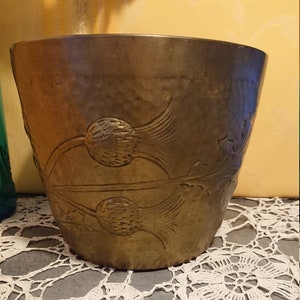 Puede incluir: Una maceta de metal martillado con un diseño de cardo. La maceta tiene una forma ancha, ligeramente acampanada y una superficie texturizada. El diseño de cardo está grabado en el metal, agregando un elemento decorativo. La maceta es de un color metálico cálido.