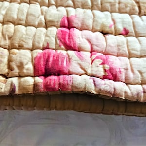 May include: A close-up of a beige quilted fabric with a floral pattern in shades of pink and white. The fabric is textured and has a slightly worn look.