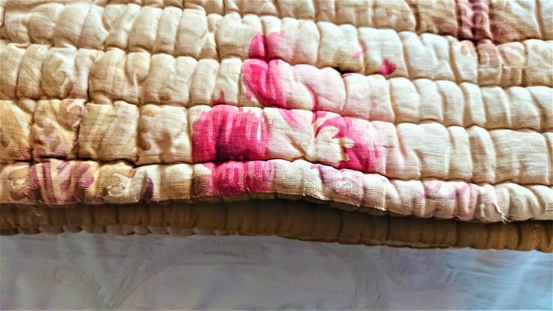 May include: A close-up of a beige quilted fabric with a floral pattern in shades of pink and white. The fabric is textured and has a slightly worn look.