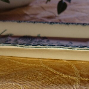 May include: A white oval ceramic dish with a blue and green floral design around the rim. The dish is on a brown fabric surface.