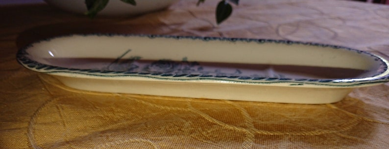 May include: A white oval ceramic dish with a blue and green floral design around the rim. The dish is on a brown fabric surface.
