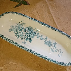 May include: A white oval ceramic dish with a blue and green floral design. The dish has a scalloped edge and three indentations.