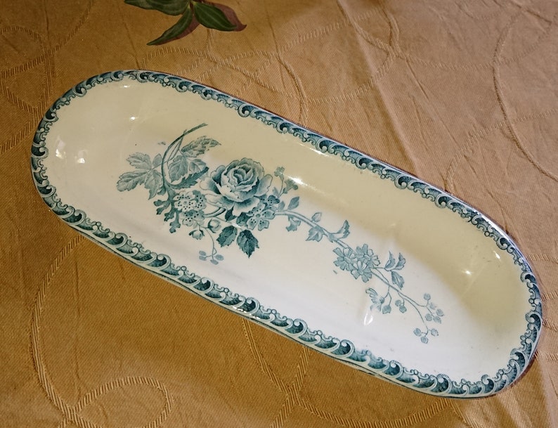 May include: A white oval ceramic dish with a blue and green floral design. The dish has a scalloped edge and three indentations.