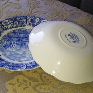 Op de afbeelding: Twee blauwe en witte keramische borden met een gegolfde rand. Het bovenste bord heeft een bloemenpatroon en een sc&egrave;ne van een stad. Het onderste bord heeft een witte achtergrond met een blauw bloemenpatroon en de tekst "Made in England".