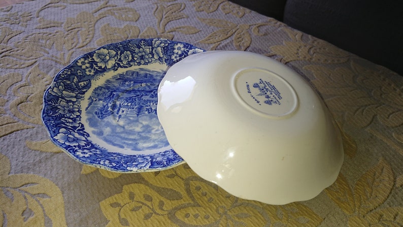 Op de afbeelding: Twee blauwe en witte keramische borden met een gegolfde rand. Het bovenste bord heeft een bloemenpatroon en een sc&egrave;ne van een stad. Het onderste bord heeft een witte achtergrond met een blauw bloemenpatroon en de tekst "Made in England".