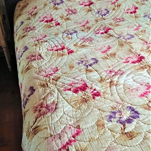 May include: A vintage floral quilt with a cream background and pink, purple, and brown floral patterns. The quilt is quilted in a circular pattern.
