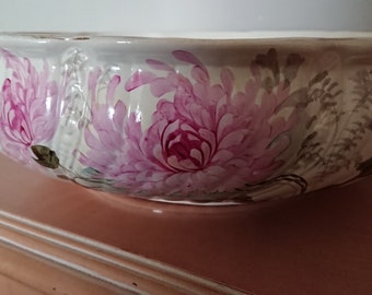 May include: A white ceramic bowl with a scalloped rim, adorned with large pink and purple floral designs. The bowl is resting on a light brown surface, showcasing its decorative floral pattern.
