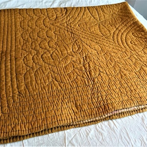 May include: A mustard yellow quilted throw blanket with a decorative pattern. The blanket is folded and lying on a white surface.