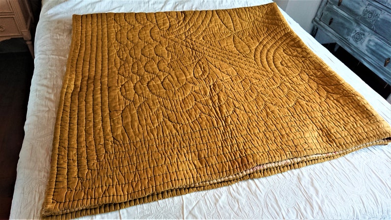 May include: A mustard yellow quilted throw blanket with a decorative pattern. The blanket is folded and lying on a white surface.