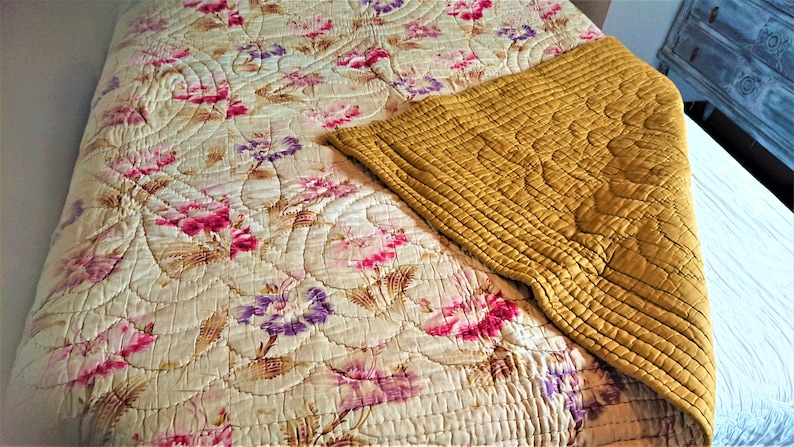 May include: A vintage floral quilt with a yellow quilted throw on top. The quilt features a floral pattern with pink, purple, and white flowers on a cream background.