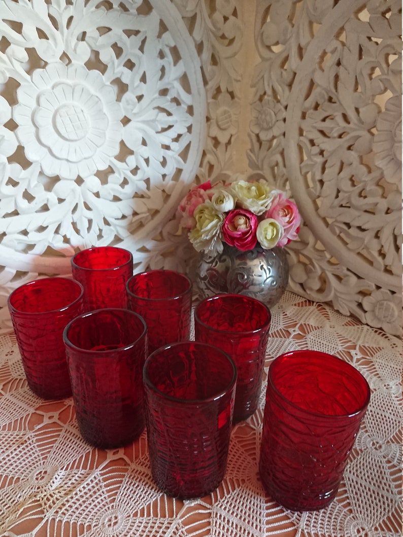 May include: Set of eight red glass tumblers with a textured surface. The glasses are arranged in a circle on a white crocheted tablecloth.