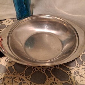 May include: A polished, oval stainless steel serving bowl with two handles. The bowl has a smooth, reflective surface and a slightly raised rim. The bowl is empty and appears clean.