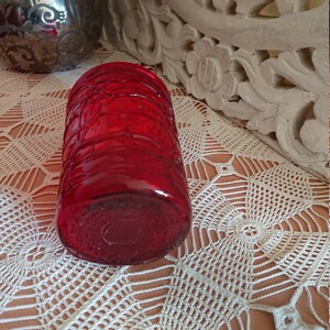 May include: A red glass candle holder with a textured design. The holder is sitting on a white crocheted surface.