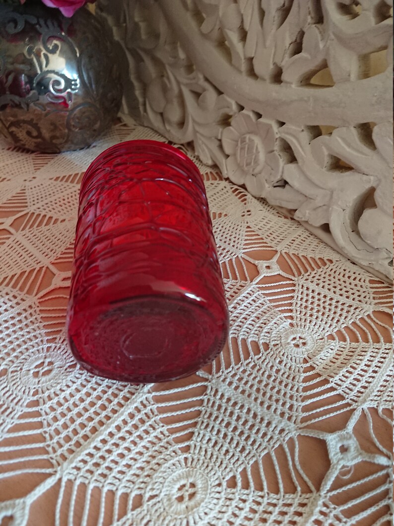 May include: A red glass candle holder with a textured design. The holder is sitting on a white crocheted surface.