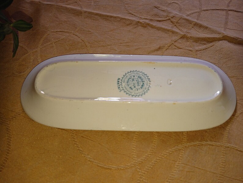 May include: A white oval ceramic dish with a green and white seal on the bottom. The seal reads "Fabrique en France" and "J. & H. NOULLIER, PARIS".